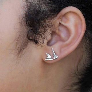 Close-up of an ear wearing silver earrings with a witch hat and broomstick design.