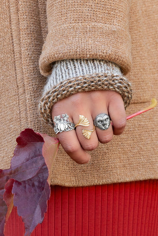 A close-up of a person's hand wearing three distinct rings, styled with a chunky knit beige sweater, a white ribbed cuff, and a red skirt, with autumn leaves in the background.