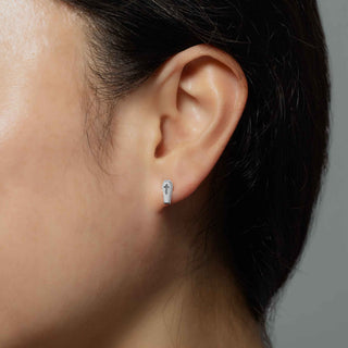 A close-up of a person's ear wearing a small silver earring shaped like a coffin with a cross on top, against a neutral gray background.