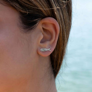 A close-up of a person's ear wearing a small silver fish stud earring, with a soft blurred outdoor background.