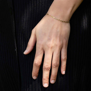 A delicate gold chain bracelet worn on a person's wrist, shown against a dark ribbed fabric background.