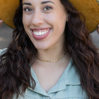A close-up lifestyle shot of a smiling woman with long wavy brown hair, wearing a delicate gold chain necklace, a light sage green shirt, and holding a mustard yellow hat.