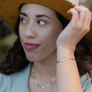 A close-up lifestyle shot of a woman wearing a delicate gold chain bracelet with , along with a gold necklace and a wide-brimmed brown hat.