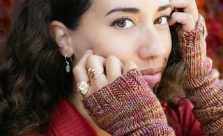 A close-up lifestyle shot of a woman wearing gold rings and a silver dangle earring, styled with a chunky knit sweater in autumnal colors.