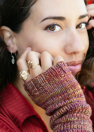 A close-up lifestyle shot of a woman wearing a chunky knit fingerless glove in a variegated blend of purple, orange, and brown tones, paired with gold rings and a red top.