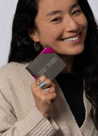 A smiling woman with long dark hair holds a gray Nina Designs box. She is wearing a beige knit cardigan over a black top, with silver rings on her fingers and a earring on her ear, against a plain light gray background.