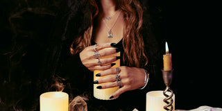 A person with long wavy brown hair holds a yellow candle, wearing multiple silver rings, a silver necklace, and a silver bracelet. The scene is dark with lit candles on a black background.