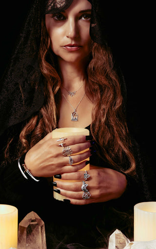 A woman in a black hood and cloak holds a glowing candle, wearing multiple silver rings and a silver pendant necklace against a dark background with candles and crystals.