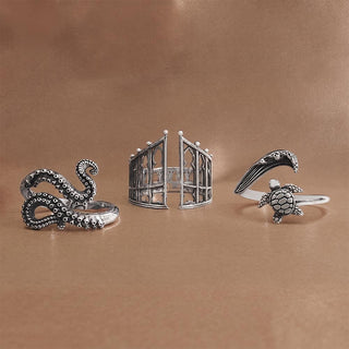 Three silver-toned rings displayed on a warm brown background: an octopus ring with raised dot detailing, a birdcage-style open ring, and a turtle ring with a wave motif.