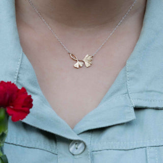 Gold necklace with leaf design worn by a person in a light blue shirt, with a red flower in the foreground.