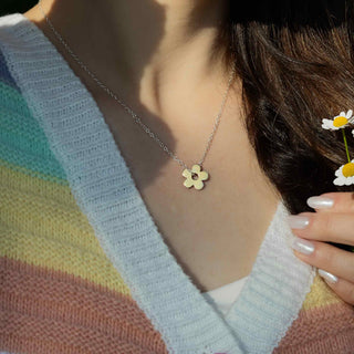 Person wearing a necklace with a yellow flower pendant, holding a small white flower.