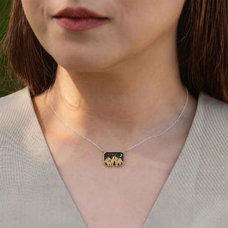 A close-up of a woman wearing a delicate silver chain necklace with a small rectangular pendant featuring a black background and a gold-toned mountain silhouette.