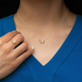 A close-up lifestyle shot of a model wearing a delicate gold and silver bird pendant necklace, paired with a matching gold ring on her finger, set against a vibrant blue V-neck top.