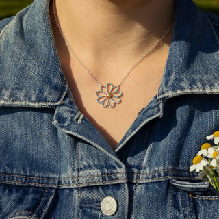 Person wearing a silver necklace with a floral pendant against a denim jacket background