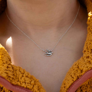 A close-up of a person wearing a delicate silver chain necklace with a small flying bird pendant, styled with a textured mustard yellow knit top.