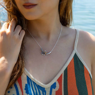 Close-up of a person wearing a silver necklace with a blue pendant, standing by a body of water.