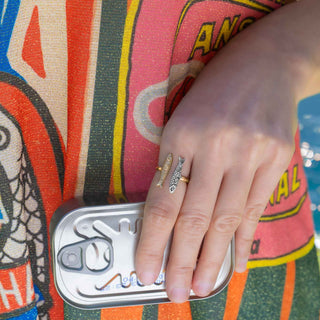 A close-up of a person's hand resting on a silver can, wearing two fish-shaped rings on the fingers. The person is wearing a colorful patterned top with orange, green, pink, and yellow stripes.