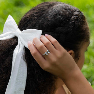 Model with dark hair with white bow in hair wearing silver bow ring. 