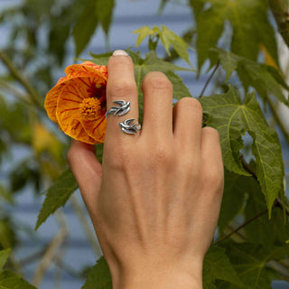 A hand wearing a silver wrap ring with two birds, posed next to a bright orange flower with green leaves in the background.