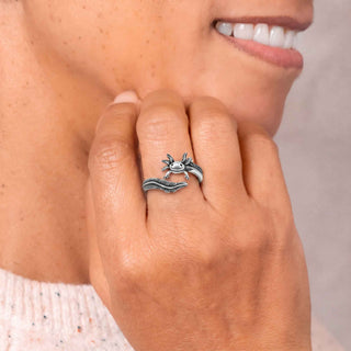 A close-up lifestyle shot of a person's hand wearing a silver axolotl ring, set against a soft neutral background.