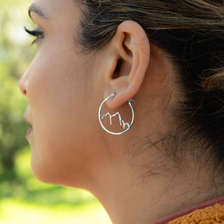 Close-up of a person wearing a silver hoop earring with a mountain design, outdoors.