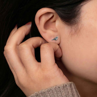 A close-up of a person's ear wearing a small silver bird stud earring, with their hand touching the earlobe