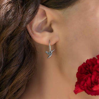 Silver hummingbird earring worn by a person with a blurred red flower in the background