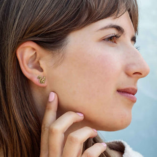 A close-up side profile of a woman wearing a small bronze squirrel stud earring . Her hand gently touches her cheek, and the background is a soft, out-of-focus light blue.