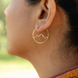 Close-up of a person wearing bronze hoop earrings with mountain design.