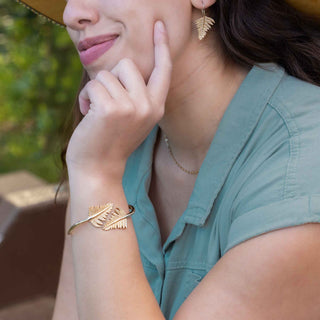 Woman wearing bronze fern earrings and bracelet, with a blurred green background