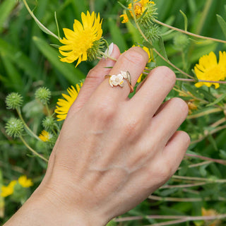 A close-up of a hand wearing a delicate bronze ring with a flower and leaf design, set against a background of green foliage and yellow wildflowers.