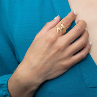 A close-up of a person's hand wearing a gold ring with a textured, organic shape, resting on their teal blue top.