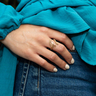 A close-up of a person's hand wearing a gold ring with a yarn and scissor ring, resting on blue denim jeans and a teal blouse.