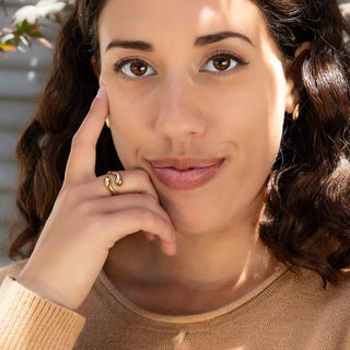 Woman with hand near her face, wearing a Bronze Adjustable Teardrop Swirl Ring and earring, with a blurred outdoor background.