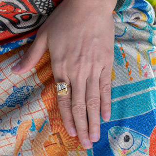 A gold ring with a square bezel set with a silver and white patterned stone, worn on a person's ring finger over a colorful patterned fabric.