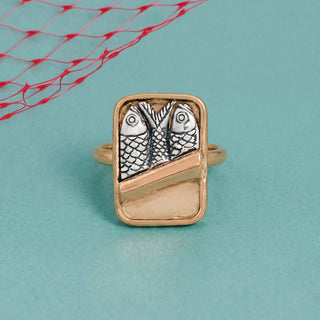 A rectangular gold ring featuring two silver fish motifs and a diagonal gold bar, displayed on a light blue background with a red net pattern in the top left corner.