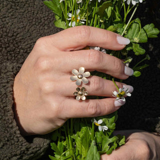 Hand wearing bronze flower-shaped ring holding a bouquet of green leaves and small white flowers.