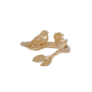 A gold-toned ring featuring a small bird perched on a branch with leaves and a flower, photographed on a plain white background.