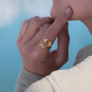 A close-up lifestyle shot of a person wearing a gold ring shaped like a bird perched on a branch, resting on their ring finger against a soft blue background.