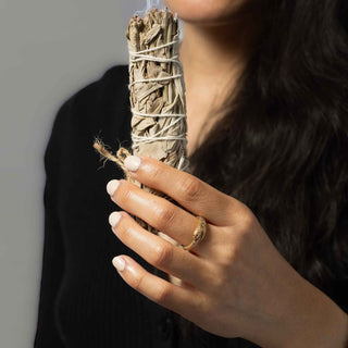 A close-up of a person holding a white sage smudge bundle while wearing a textured bronze snake ring on their hand.