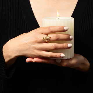 A close-up of a person in a black ribbed top holding a lit cream-colored candle, with a gold ring on one finger.