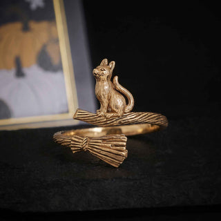 A gold-toned open ring featuring a sculpted cat sitting on a broom, displayed on a dark textured surface with a blurred framed picture in the background.