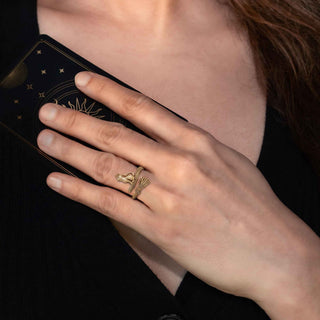 A close-up of a person's hand wearing a gold ring with a cat, resting on a black tarot card with gold celestial illustrations.