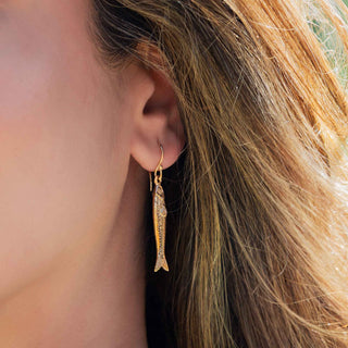 A close-up of a person's ear wearing a small gold fish-shaped dangle earring, with light brown hair visible against a softly blurred outdoor background.