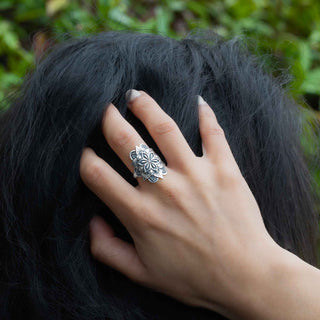 A close-up lifestyle shot of a hand with silver manicured nails wearing a detailed silver ring, resting on a person's dark hair against a blurred green background.